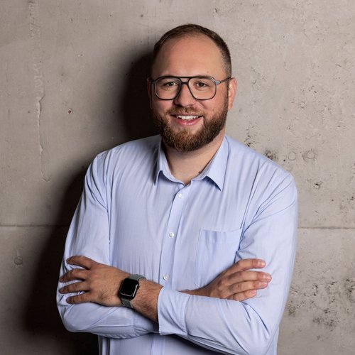 Mann mit Bart, Brille und hellblauem Hemd steht lächelnd mit verschränkten Armen vor Betonwand.